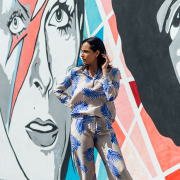 photo of woman in outfit of blouse and pants with palm tree print in front of a painted wall mural featuring David Bowie as Ziggy Stardust