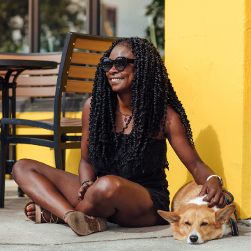 photo of woman in summer clothes wearing sunglasses and sitting outside a shop petting a small corgi dog