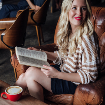 photo of smiling woman sitting in a brown leather chair reading a book in front of a table with a latte