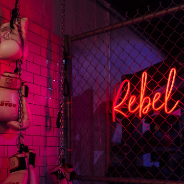 photo of wall covered in hanging pairs of boxing gloves in front of a chain link fence with a red neon script sign that reads, Rebel