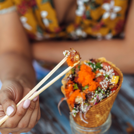 closeup photo of fish between chopsticks above a waffle cone filled with poke