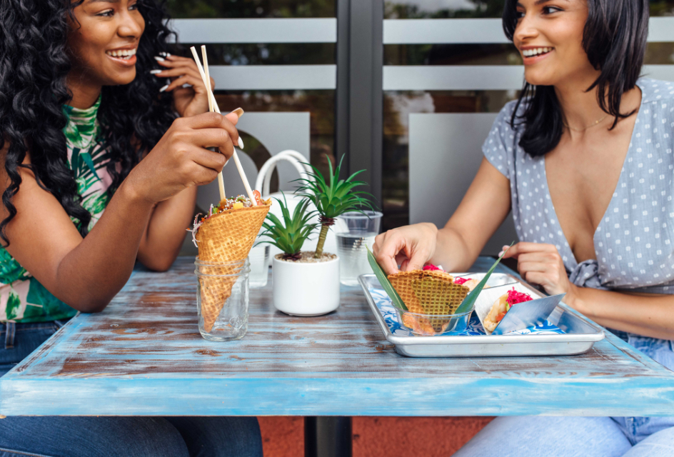 photo of two smiling women eating sushi out of waffle cones sitting at a table